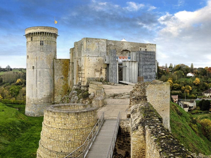 Vue du château médiéval de Falaise, avec une tour circulaire et des murs en pierre, surplombant un paysage vallonné.