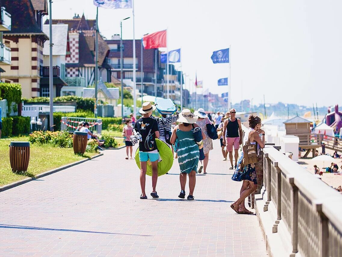 Promenade animée au bord de la plage de Cabourg, avec des touristes marchant sous le soleil et des maisons en arrière-plan.