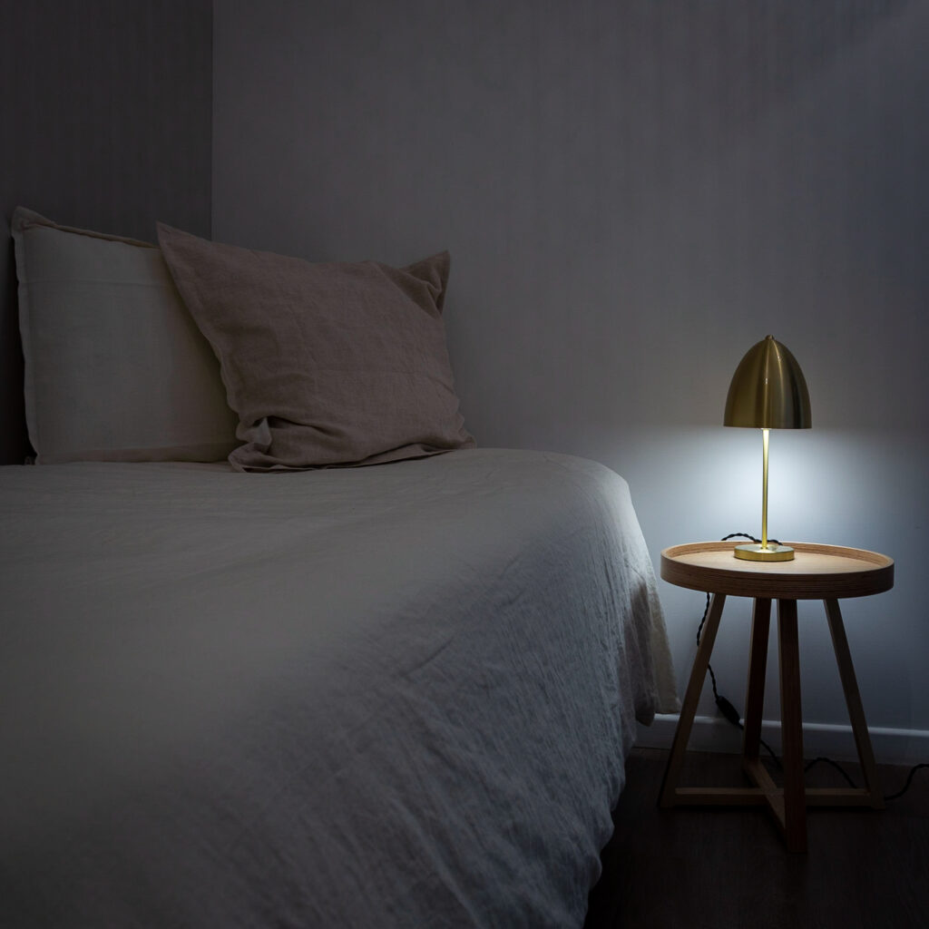 Chambre moderne avec lit, coussins et lampe dorée sur table en bois. Ambiance douce et confortable.