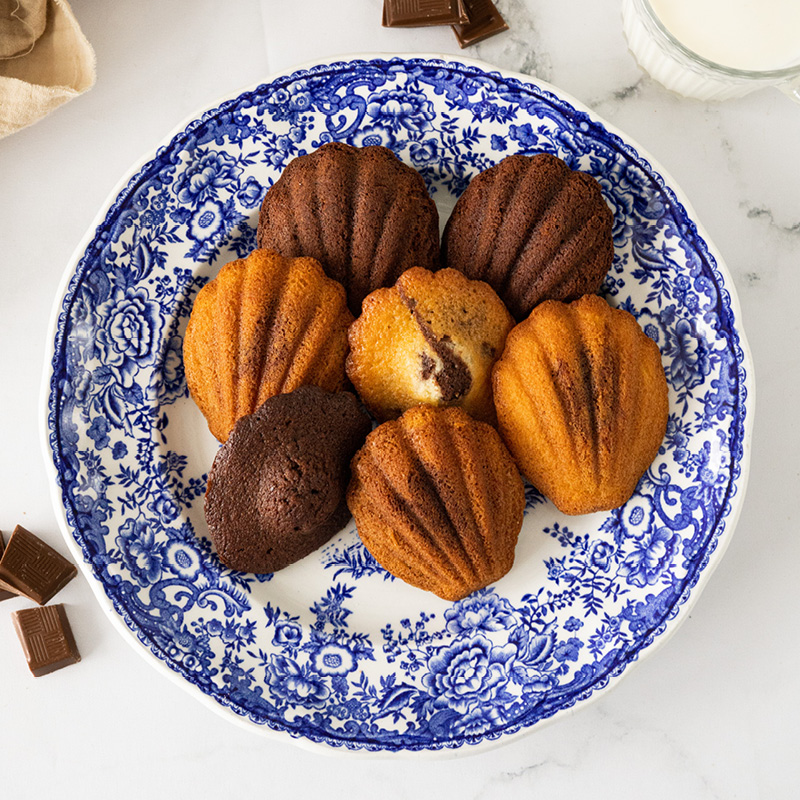 Assiette de madeleines dorées et chocolatées, présentées avec des carrés de chocolat et un verre de lait.