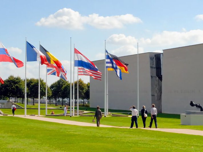 Visiteurs marchant sur un gazon vert, devant des drapeaux de différents pays et un bâtiment moderne.