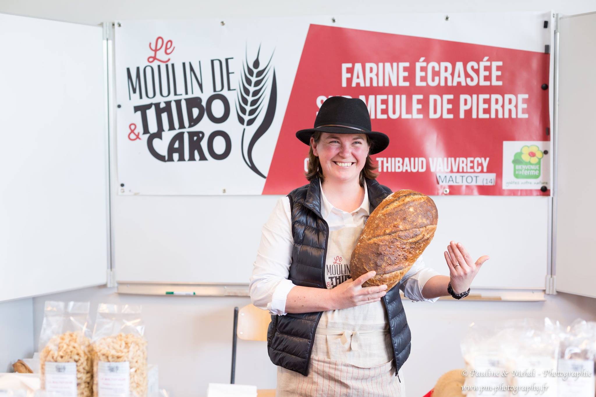 Femme souriante tenant du pain devant un stand au Moulin de Thibo et Caro, avec une enseigne en arrière-plan.