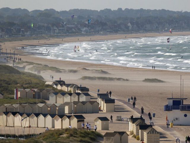 Plage de Ouistreham avec cabines, vagues et kitesurfeurs, vue ensoleillée, ambiance détendue.