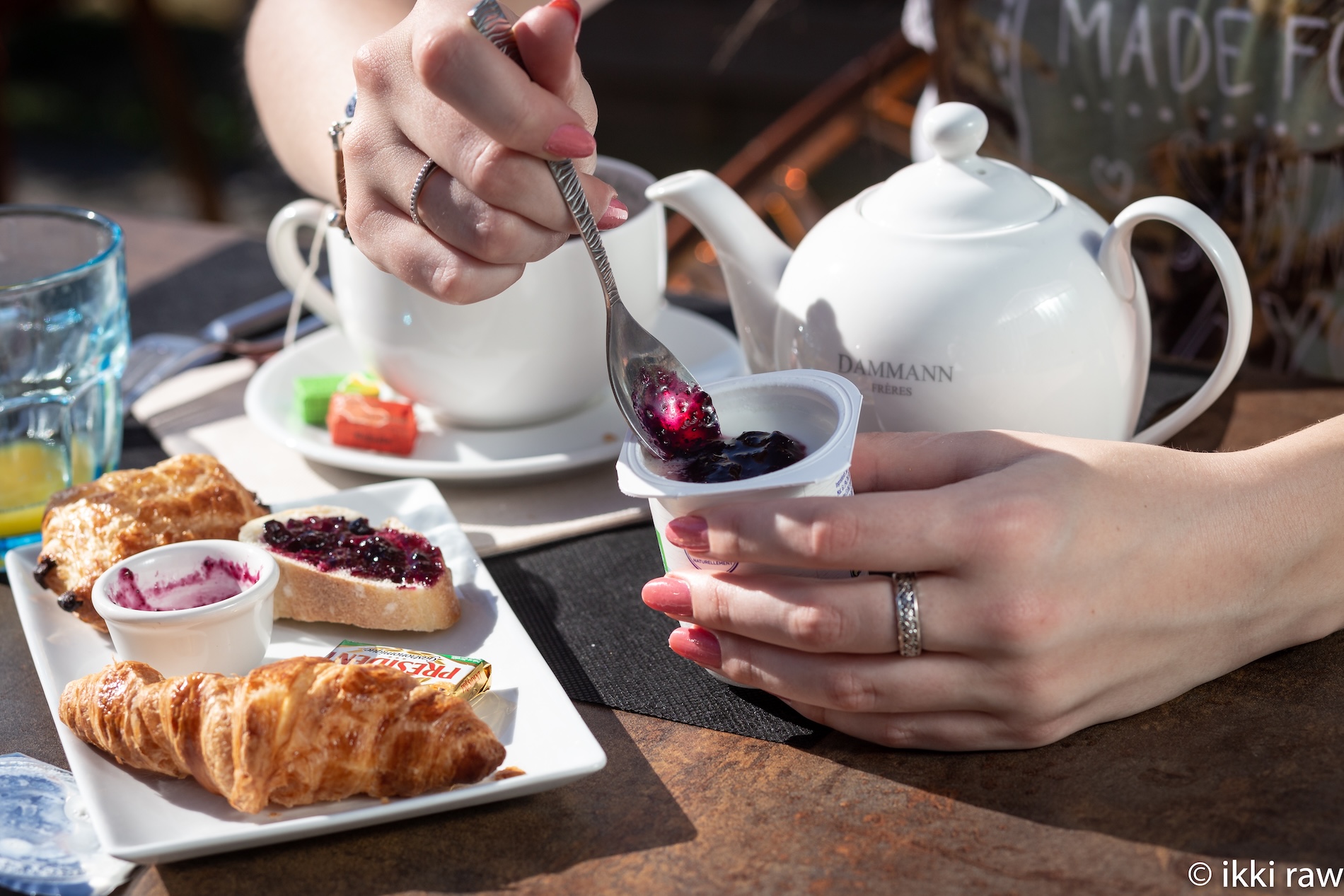 Main tenant une cuillère, se préparant à déguster un yaourt avec confiture, entouré de viennoiseries et d'une tasse de thé.